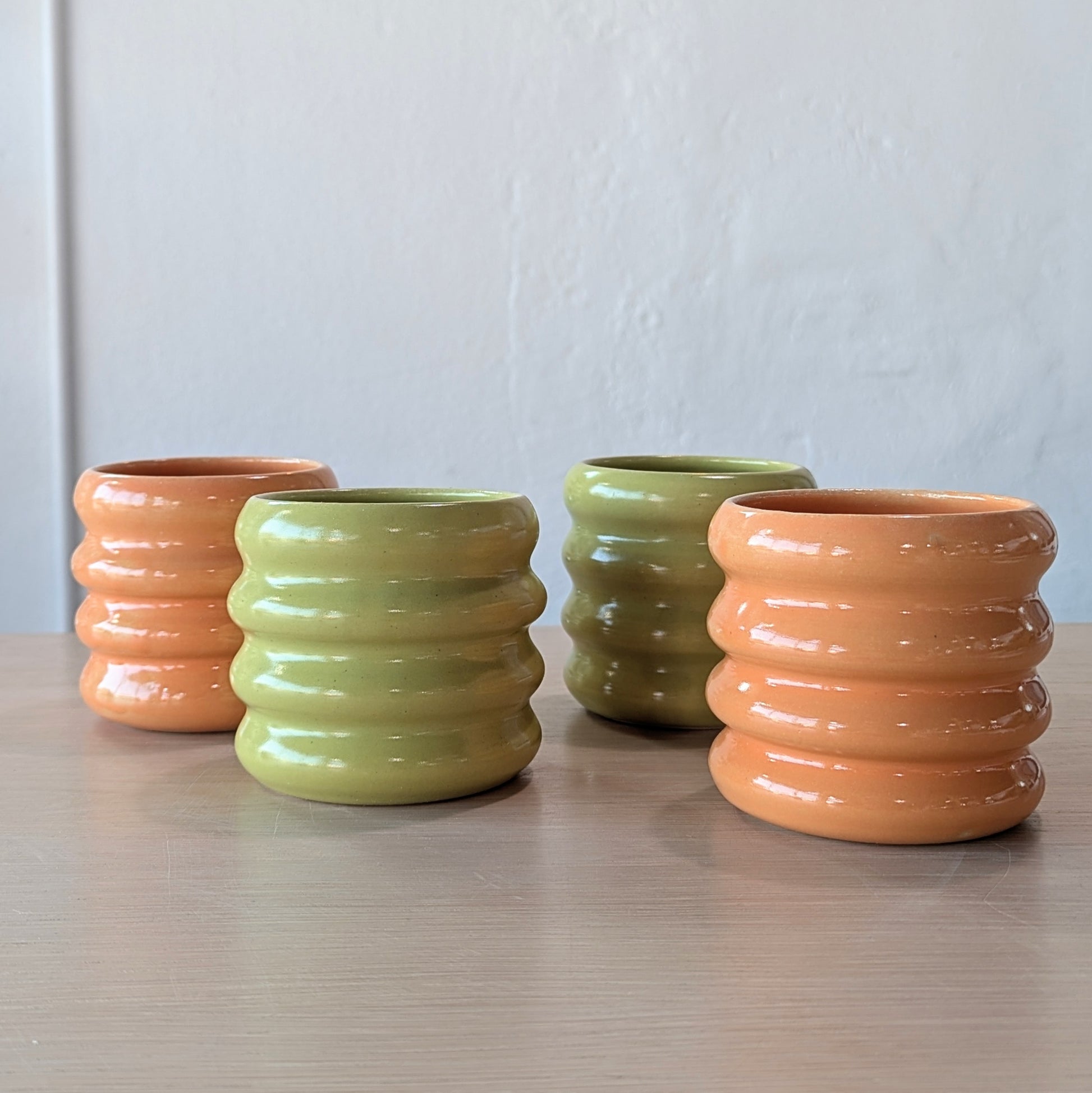 Four ribbed ceramic tumblers in green and orange on a wooden surface with a white wall background.