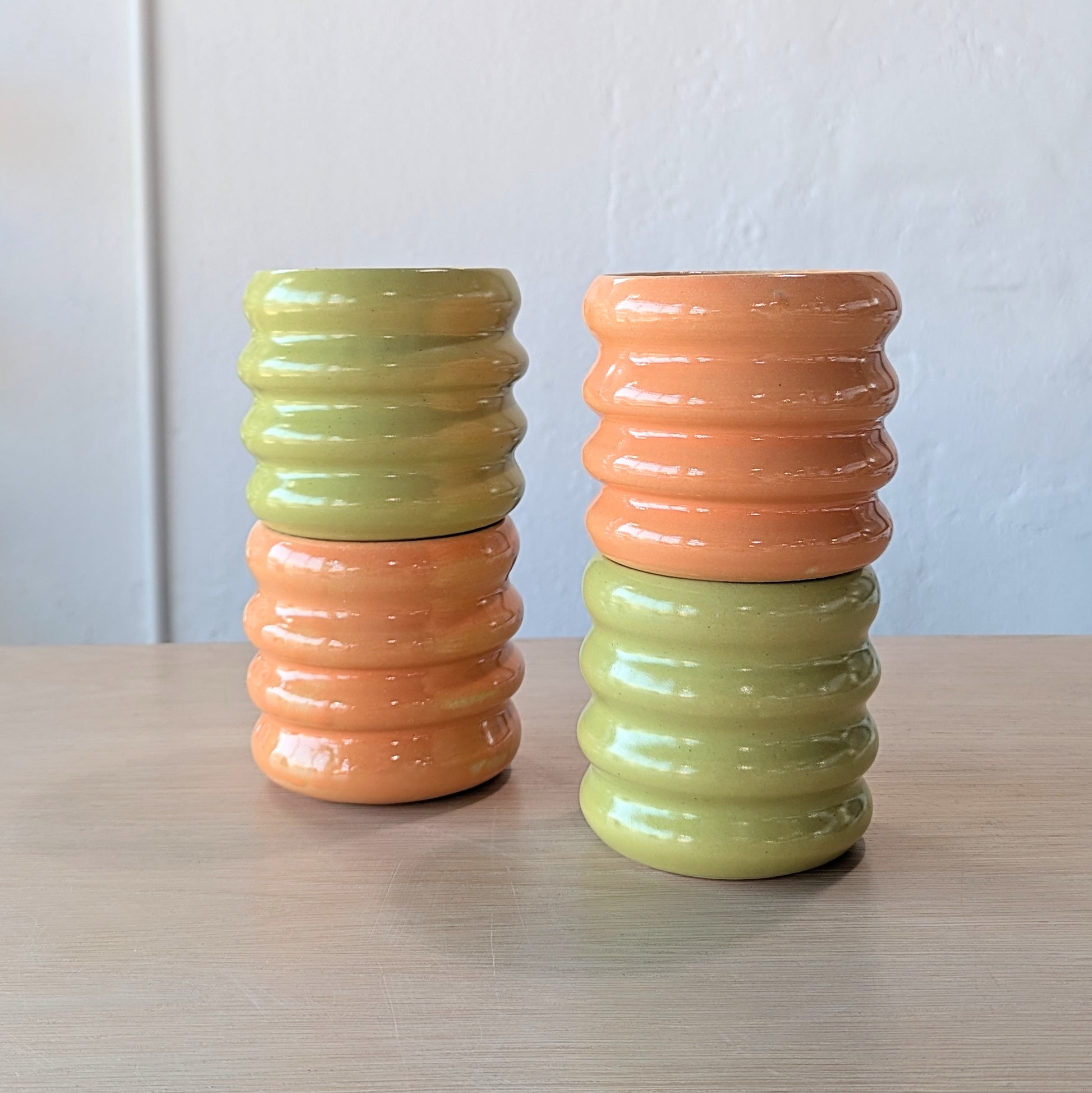 Four ribbed ceramic tumblers in green and orange on a wooden surface with a white wall background.