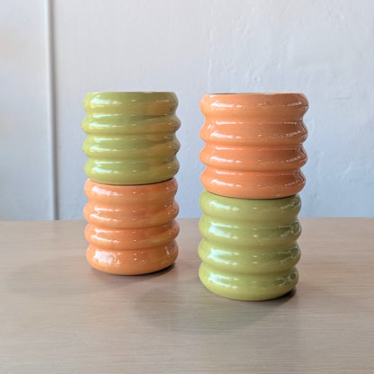Four ribbed ceramic tumblers in green and orange on a wooden surface with a white wall background.