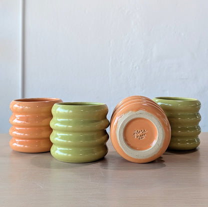 Four ribbed ceramic tumblers in green and orange on a wooden surface with a white wall background.